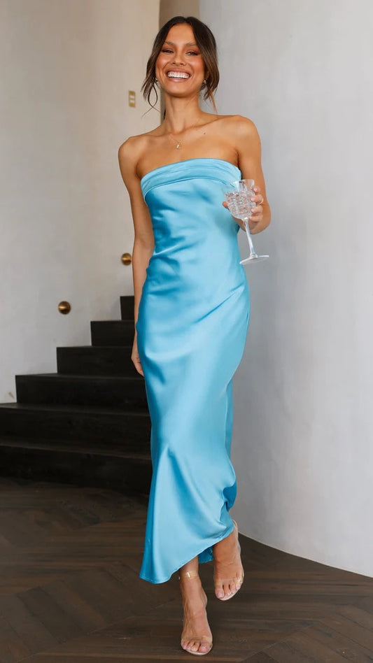 Model standing by a staircase, smiling at the camera while holding a wine glass and wearing a blue Jenna Backless Dress, a luxurious satin maxi dress with a strapless neckline and cutout elements, perfect for formal occasions